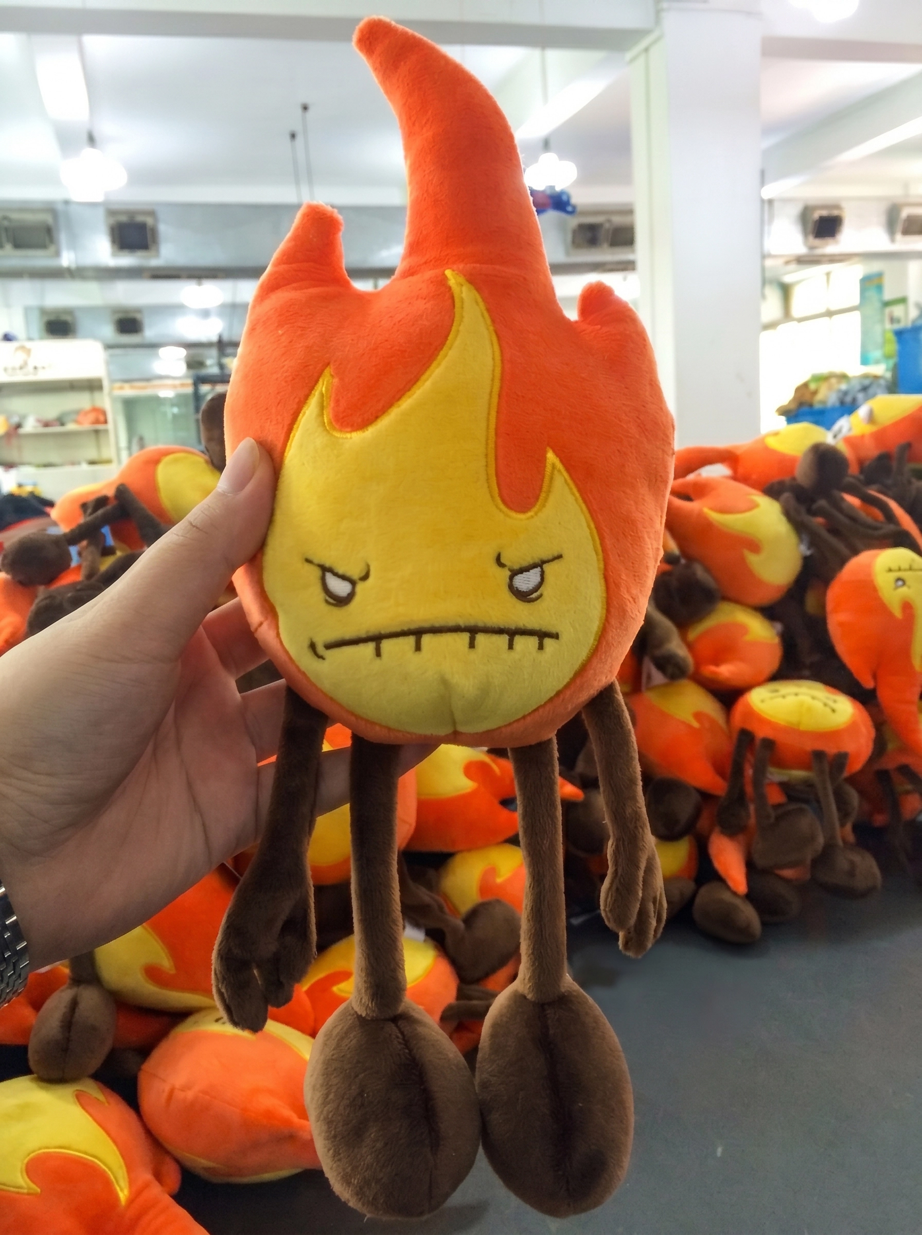 A hand holding a Fire Element plush toy showing the high-quality embroidery and long limbs, with a pile of plushies in the background during the Fire Element plush production phase.