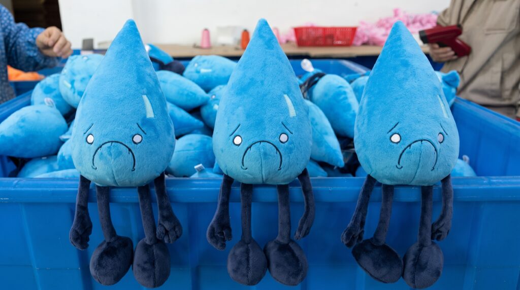 Three blue Water Element plush toys with sad expressions sitting on the edge of a blue plastic production bin filled with more plushies.