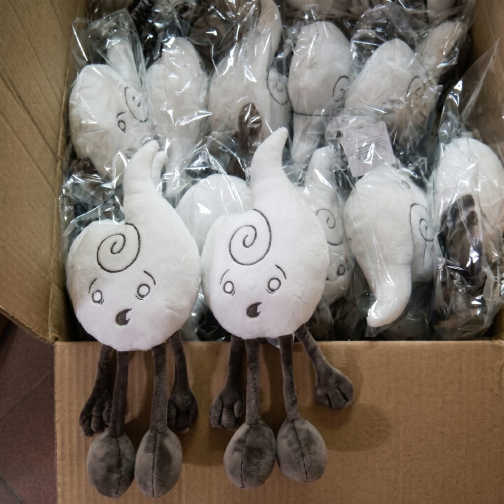 Two white Air element plush toys with swirl designs and surprised expressions sitting on a cardboard box filled with more plushies individually wrapped in plastic.
