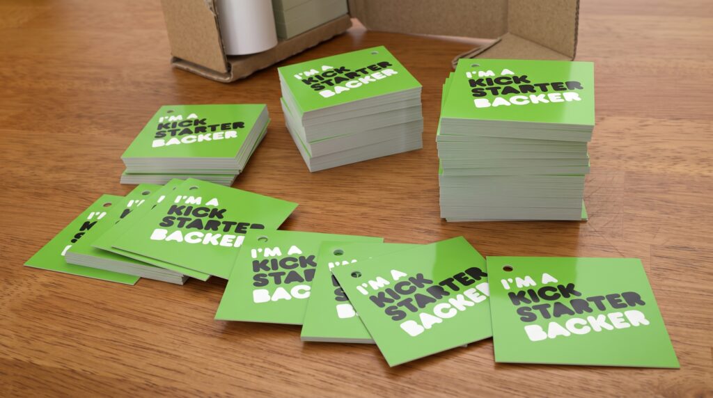 Stacks of glossy green "I'm a Backer" tags for the Elemental Plush project, shown on a wooden table as they are prepared for manual installation.