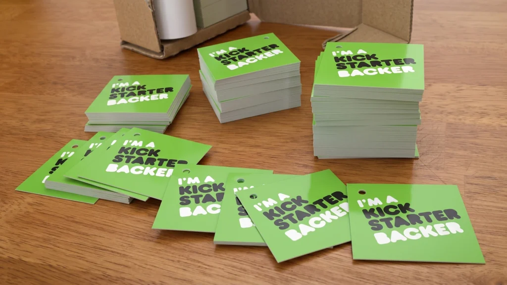 Stacks of glossy green "I'm a Backer" tags for the Elemental Plush project, shown on a wooden table as they are prepared for manual installation.