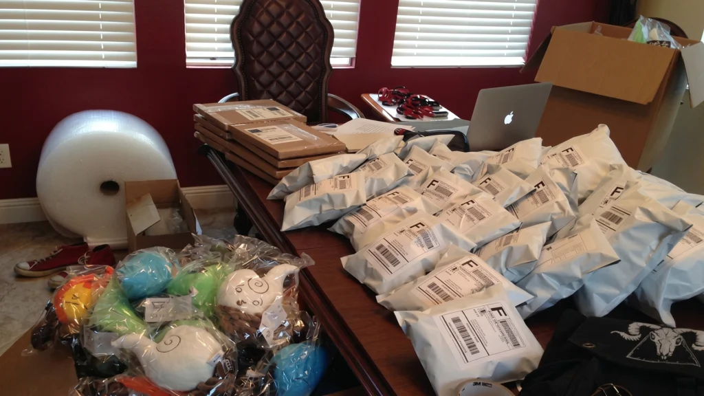 A large wooden desk covered in white bubble mailers and shipping boxes, with piles of Elemental Plush toys and a roll of bubble wrap in the background.