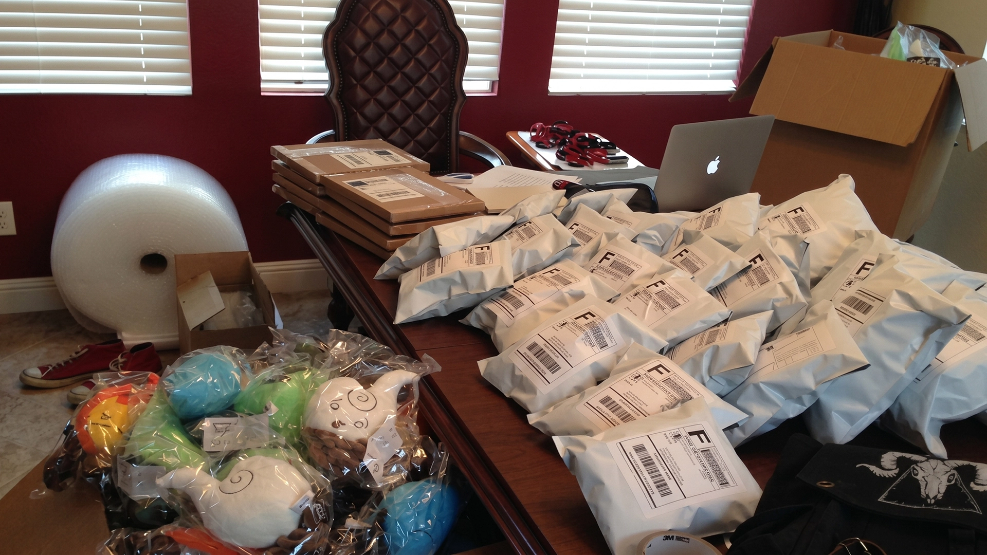 A large wooden desk covered in white bubble mailers and shipping boxes, with piles of Elemental Plush toys and a roll of bubble wrap in the background.
