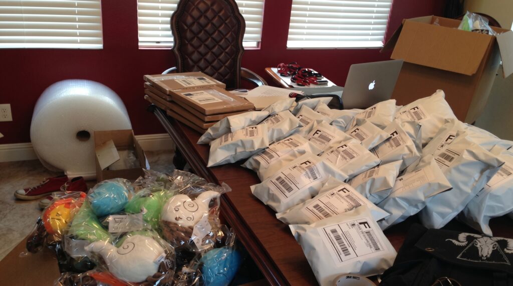 A large wooden desk covered in white bubble mailers and shipping boxes, with piles of Elemental Plush toys and a roll of bubble wrap in the background.