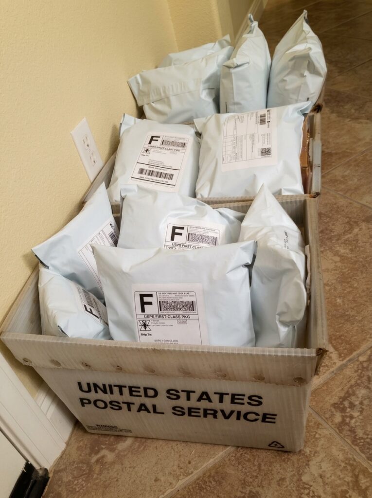 A white United States Postal Service bin sitting on a tile floor, filled to the brim with white bubble mailers addressed and ready for the first wave of shipping.