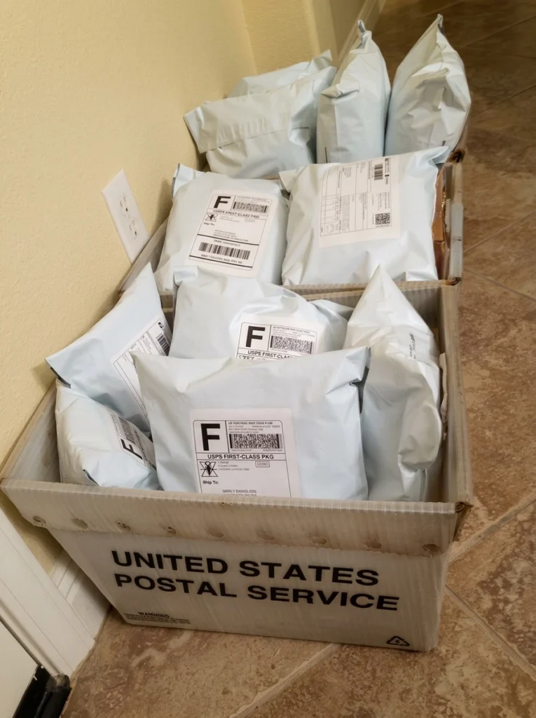 A white United States Postal Service bin sitting on a tile floor, filled to the brim with white bubble mailers addressed and ready for the first wave of shipping.